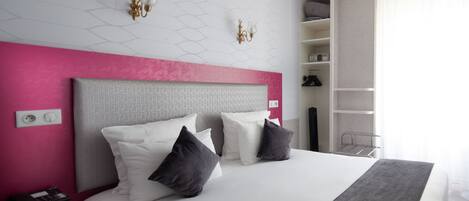 Una habitación de hotel moderna con una cama grande, una pared de acento rosa, un cabecero blanco y un armario empotrado con estantes.