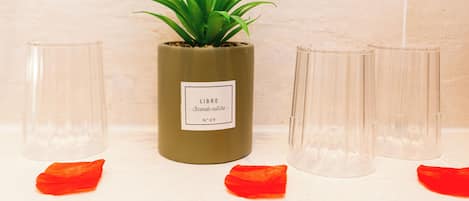 Una planta en maceta dentro de un contenedor cilíndrico con una etiqueta, rodeada de cuatro jarrones de vidrio transparente sobre una superficie de azulejos.