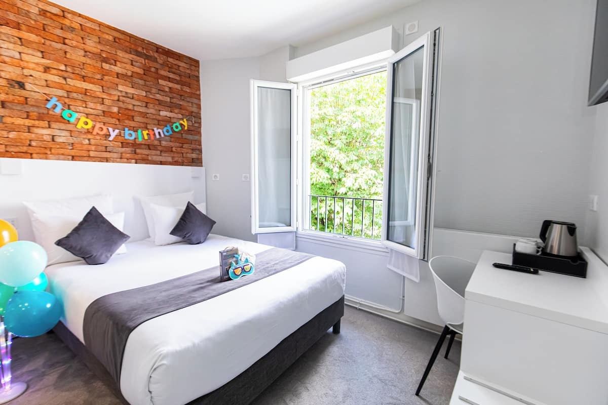 Une chambre d’hôtel moderne avec un mur d’accent en briques, un grand lit avec des oreillers, une table de chevet avec une bouilloire et une vue sur la verdure par une fenêtre ouverte.