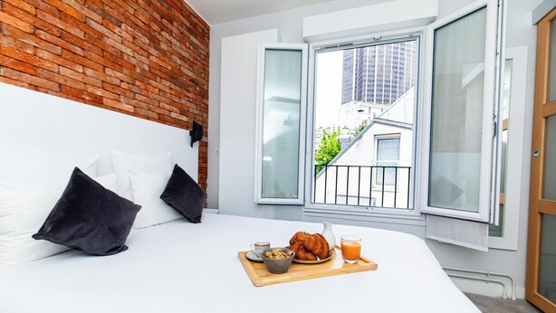 Une chambre avec un mur en briques, un lit avec une literie blanche, deux oreillers noirs, un plateau de petit-déjeuner avec des croissants, un bol de céréales et un verre de jus d’orange.