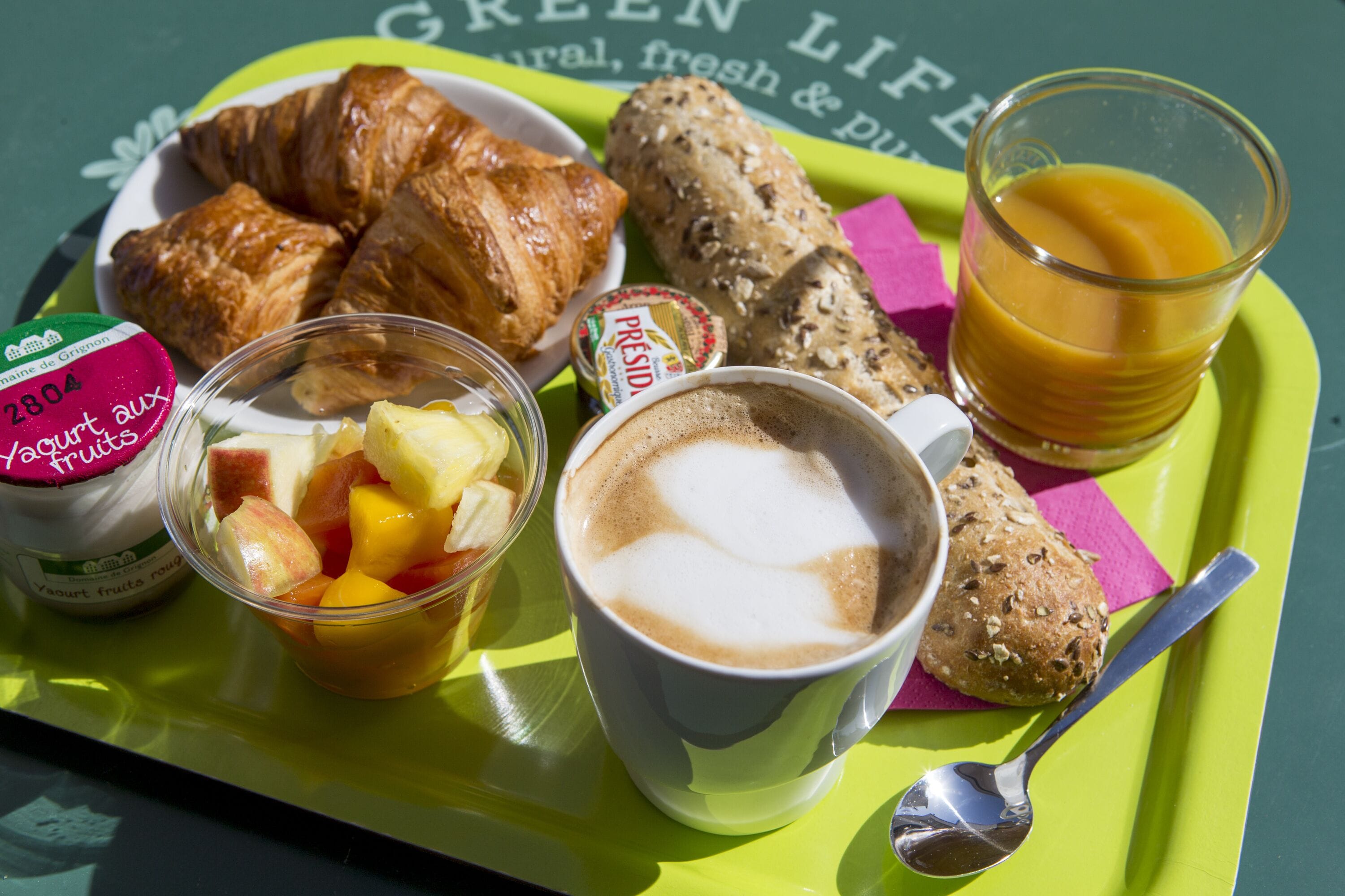 Bandeja de café da manhã com croissants, uma xícara de café, um copo de suco de laranja, uma tigela de frutas e um pote de iogurte.