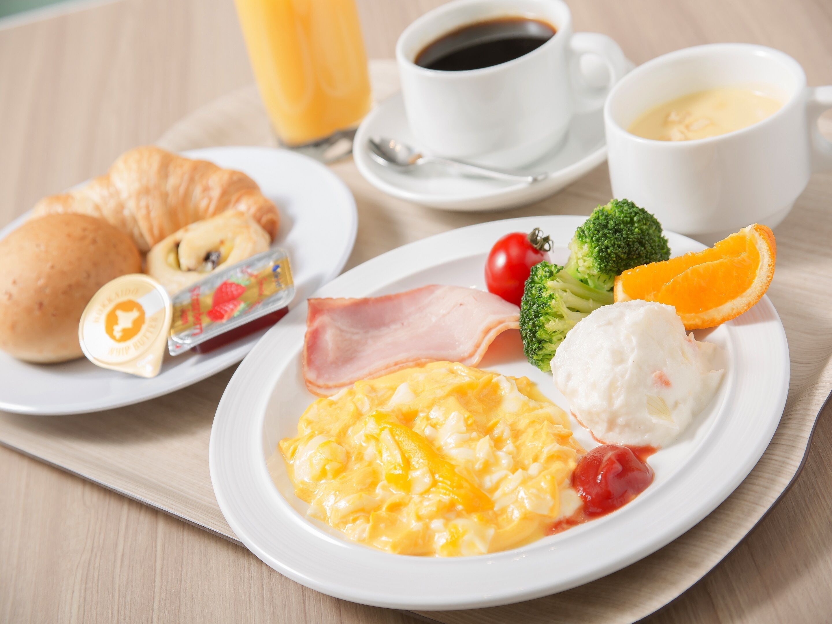 Bandeja de desayuno con cruasanes, jamón, huevos revueltos, tomate, brócoli, una rodaja de naranja y dos tazas de café.
