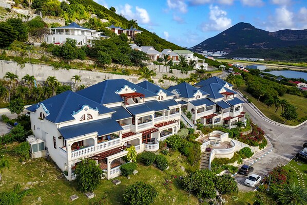 A large, multi-story hotel with blue roofs and white walls, surrounded by lush greenery and a mountainous backdrop. - Hotel L'Esplanade (Grand Case)