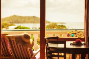 A view from a balcony overlooking a body of water with a distant island, a straw hat on a chair, and a table with drinks. - Hotel L'Esplanade (Grand Case)