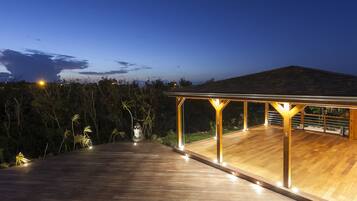 Una terraza de madera con zona cubierta, rodeada de vegetación y un cielo despejado.