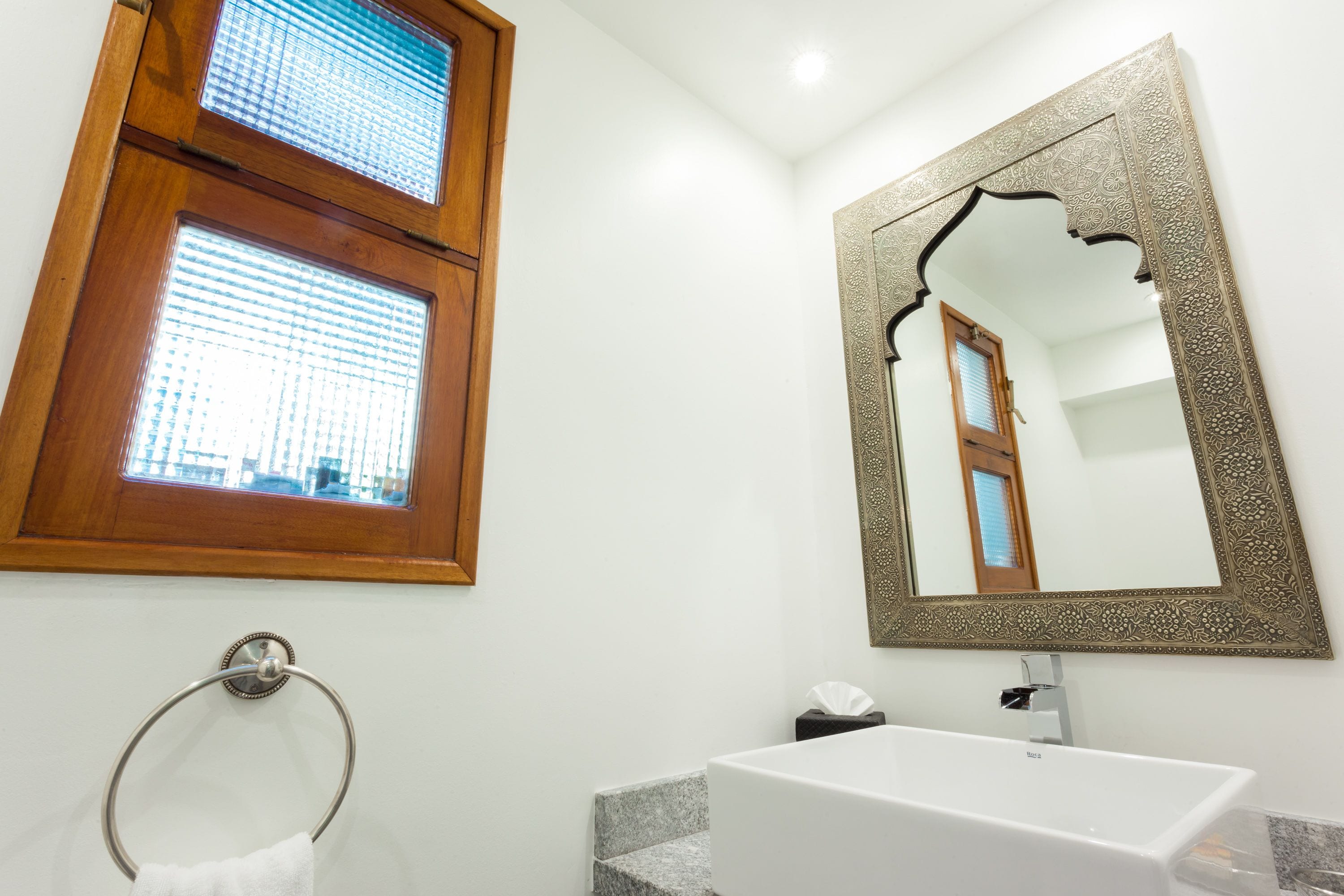 A bathroom with a wooden-framed window, a large ornate mirror, and a white sink.