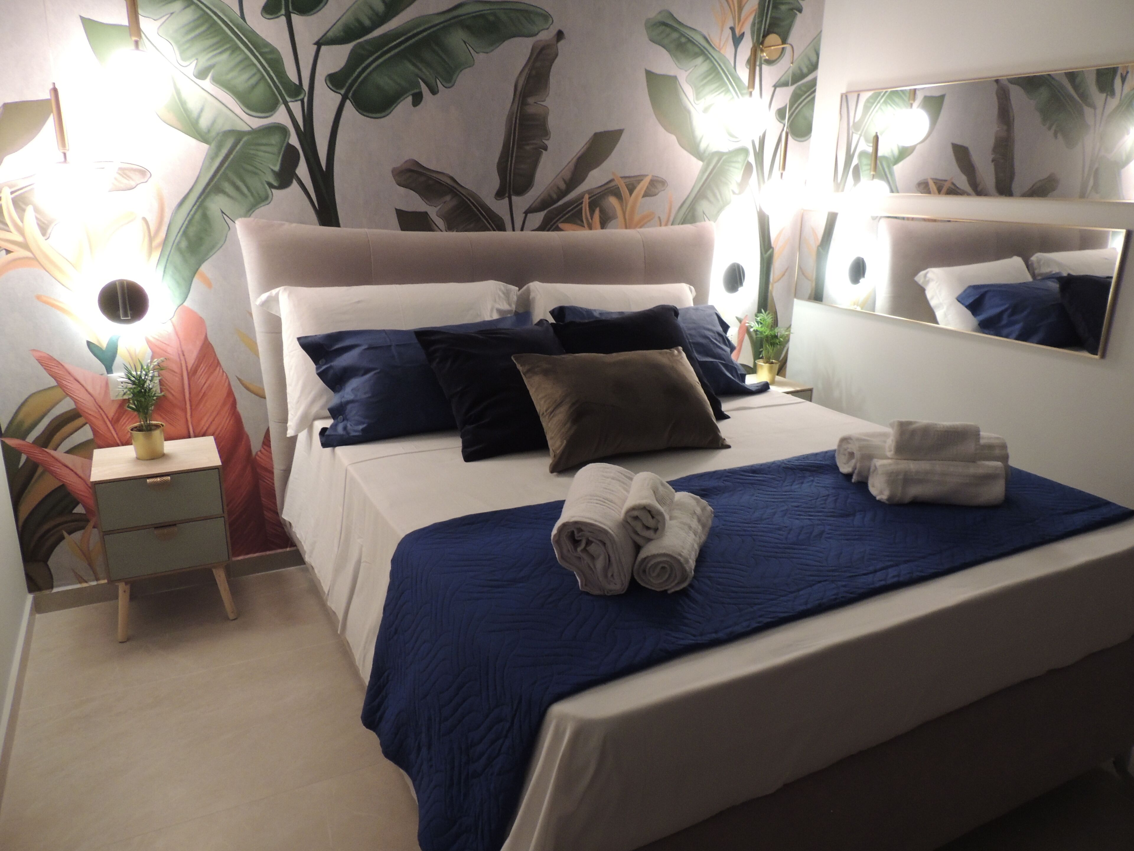 A neatly made bed with blue bedding, a bedside table with a lamp, and a large mirror.