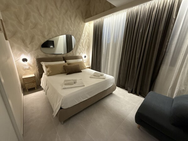 A modern bedroom with a large bed, a bedside table, a mirror, and a dark-colored armchair.