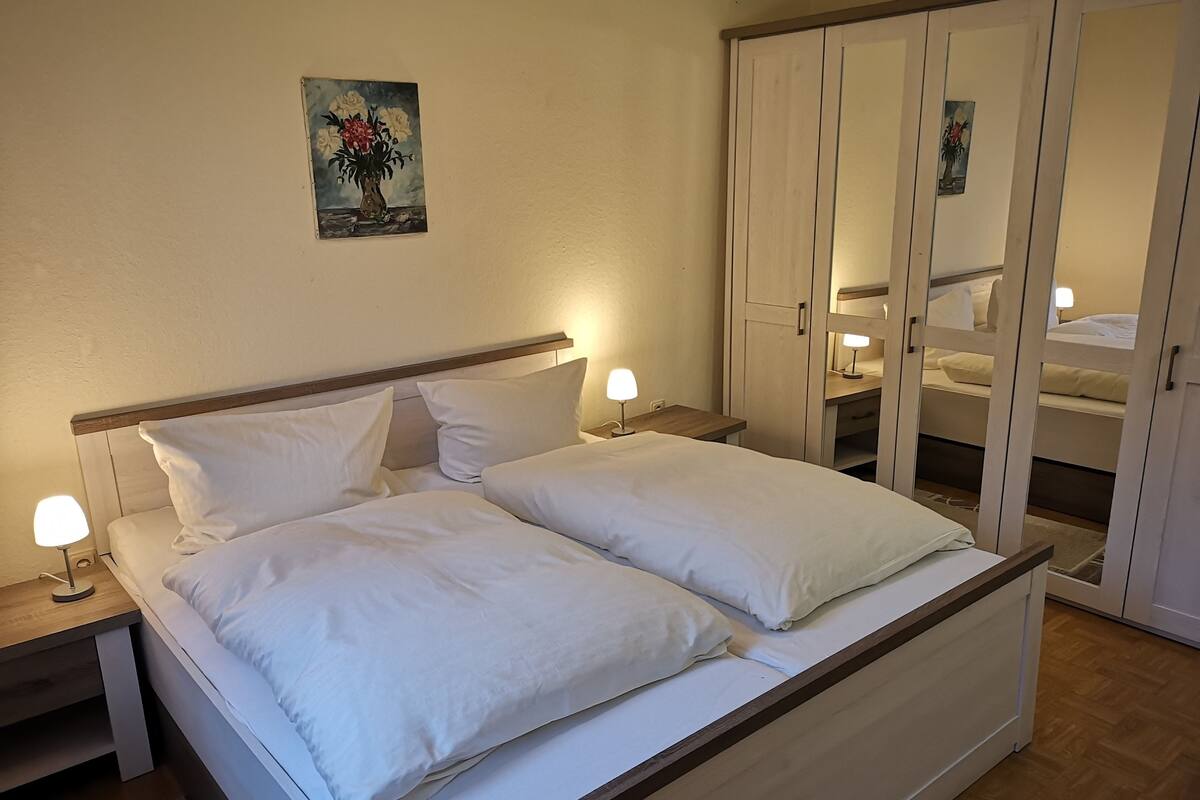 A double bed with white linens, two bedside tables with lamps, a wooden headboard, a painting on the wall, and a wardrobe with mirrors.