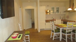 A compact kitchen with white cabinets, a wooden dining table with four chairs, and a small seating area with a chair and a table. - Ennenhof (Schneverdingen)