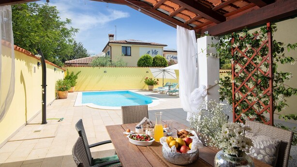 Une terrasse avec une piscine, une table à manger où l’on peut prendre le petit-déjeuner, et une vue sur une maison.