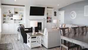 Ein Wohnzimmer mit Kamin, Fernseher, Esstisch mit Stühlen und Bücherregal mit Dekoration.