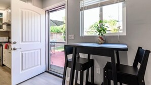 Una cocina con armarios blancos, una puerta blanca, un ventanal con persianas y una mesa con sillas.