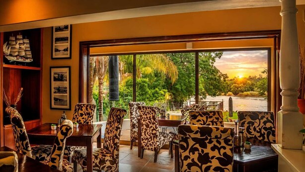 An indoor dining area with patterned chairs and a view of a sunset through large windows. - Moonriver Guesthouse (Upington)