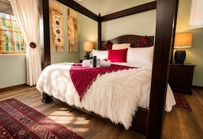 A four-poster bed with a white and red blanket, a nightstand with a lamp, and a window with curtains. - Moonriver Guesthouse (Upington)