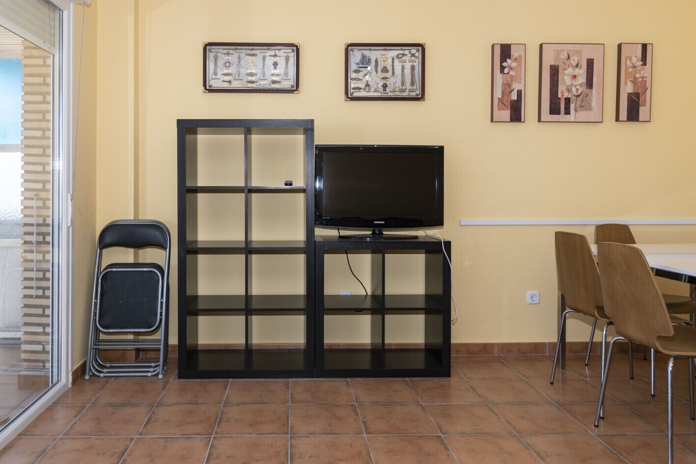 Une pièce avec un téléviseur posé sur un meuble noir, une table à manger avec des chaises, une chaise pliante et trois œuvres d’art encadrées accrochées au mur.
