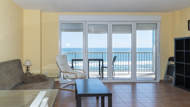 A living room with a sofa, a table, and a chair, a balcony with a view of the ocean, and a bookshelf. - FRONTALMAR, ESC-B, 8º-32 (Cullera)