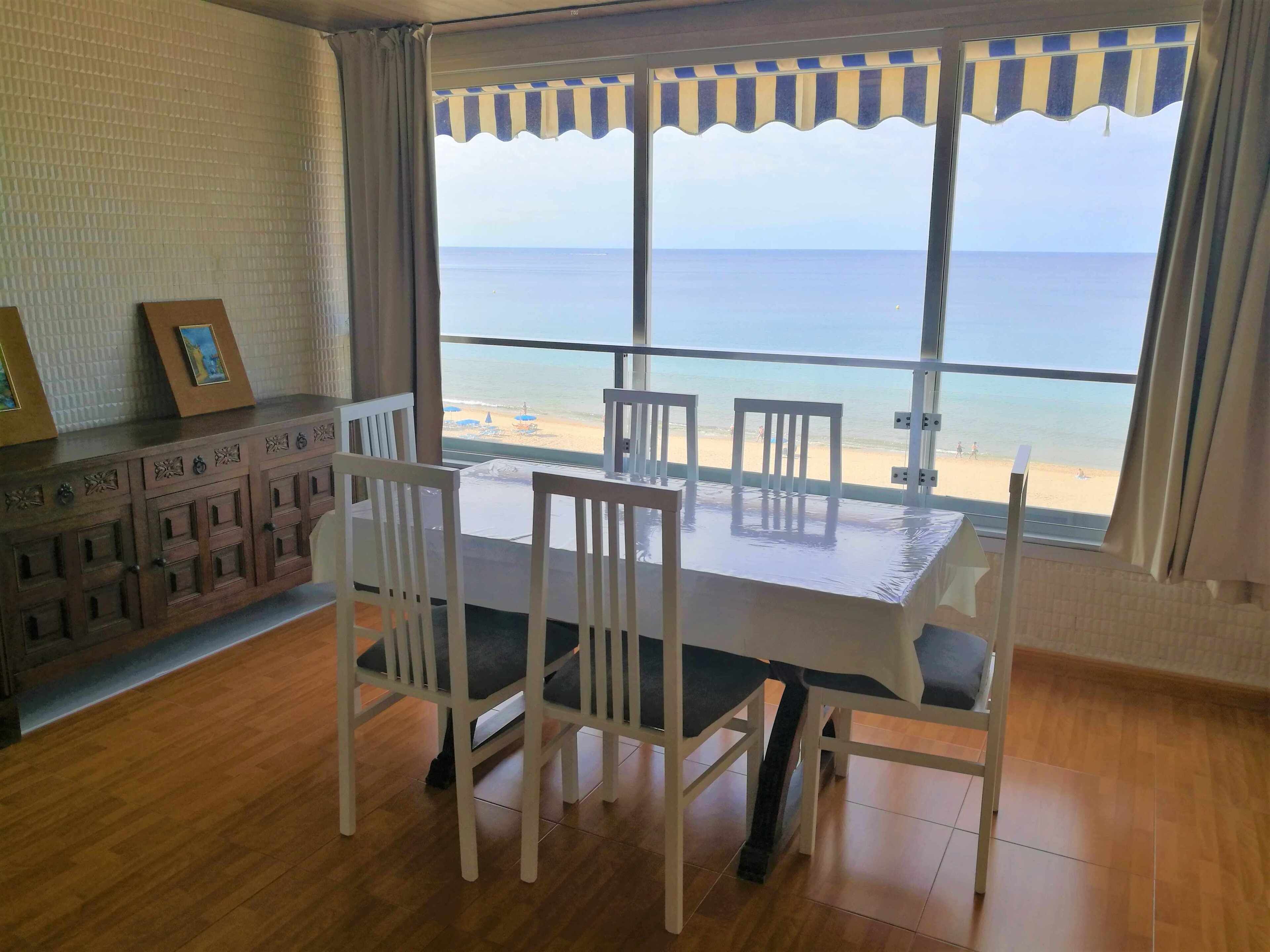 A room with a large window overlooking a beach, a wooden cabinet with decorative items, and a table with chairs.