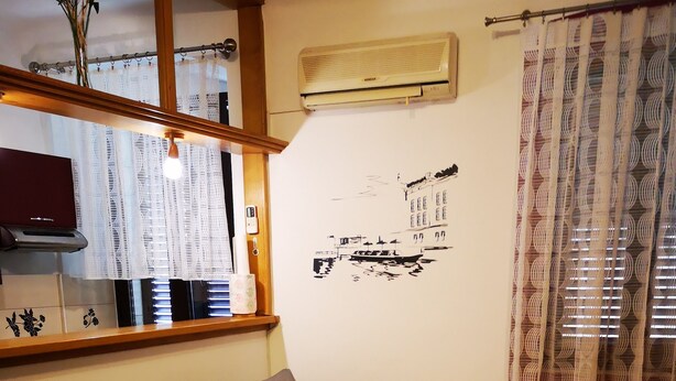 Una sala de estar con un sofá, una mesa de centro de vidrio con camino de mesa de encaje, un aire acondicionado montado en la pared y una ventana con cortinas estampadas.