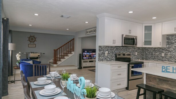 A modern kitchen with white cabinets, a microwave, and a stove. There is a dining area with a table set for four, blue chairs, and a blue accent wall. - Home away from home! (South Pasadena)