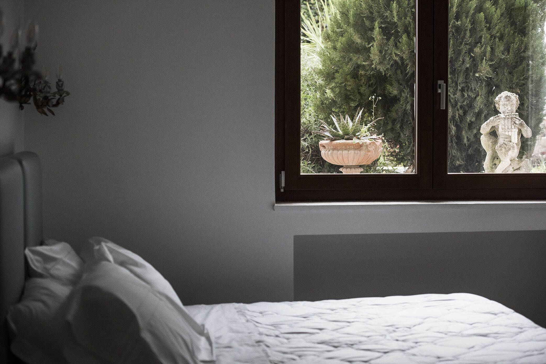 Un dormitorio con cama, ventana con vista a follaje y una estatua, además de una planta colgante.