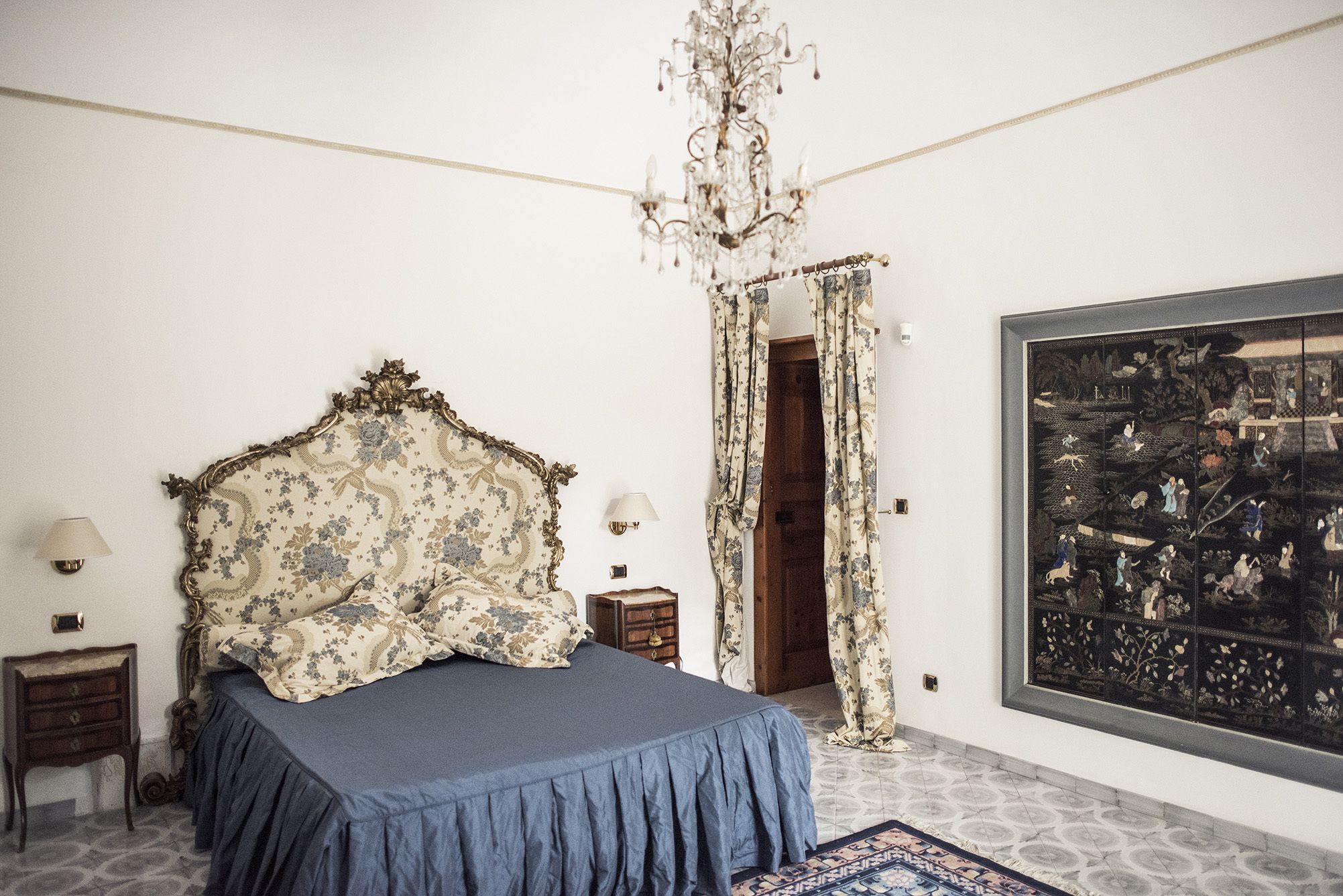 A bedroom with a large bed, a chandelier, a nightstand, a lamp, and a large painting on the wall.