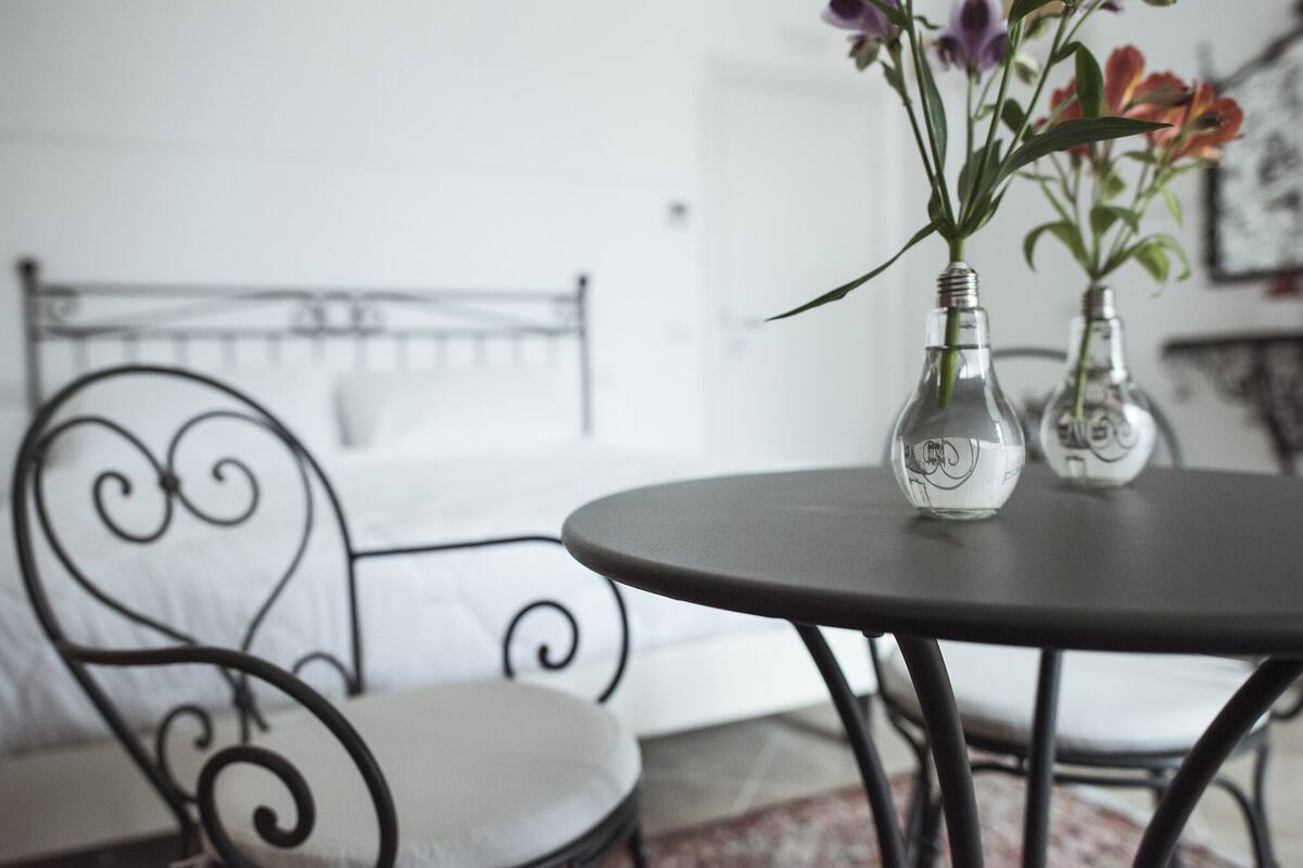 A bed with a metal headboard, a round table with two glass vases containing flowers, and a chair with a white cushion.