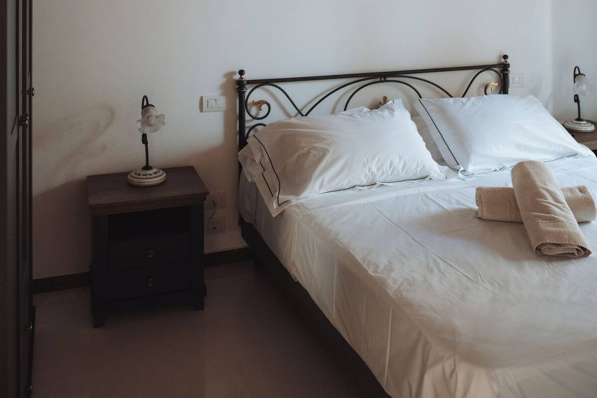 A bedroom with a wooden ceiling, a metal bed frame, white bedding, a bedside table with a lamp, and a wall-mounted light fixture.