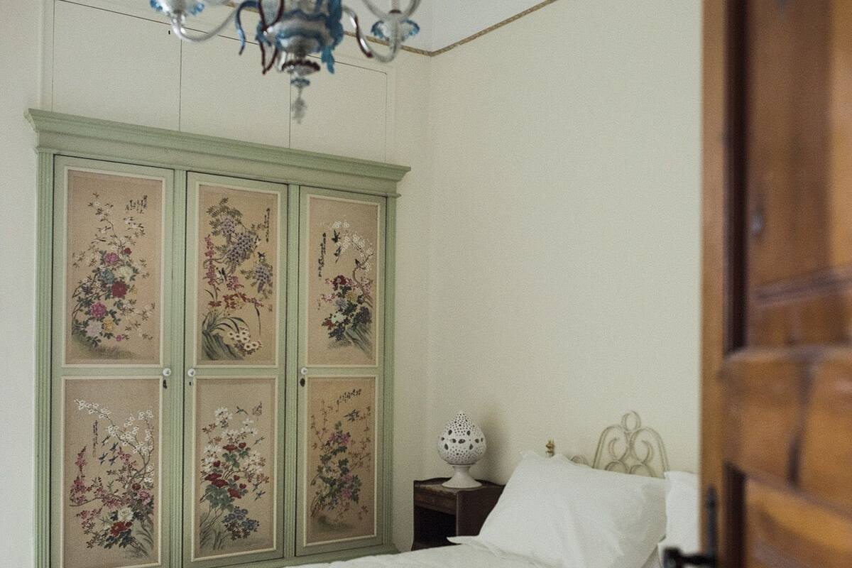 A bedroom with a bed, a nightstand, a decorative cabinet with floral panels, and a chandelier.