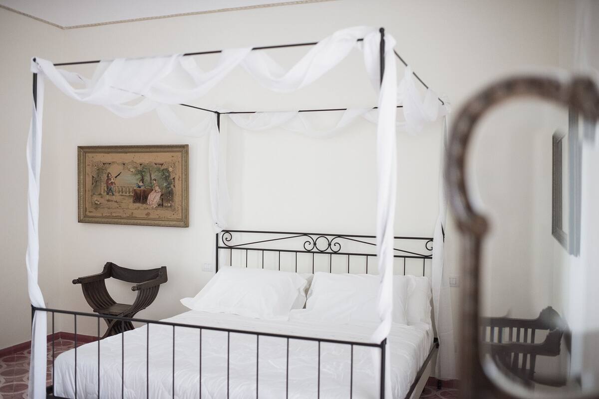 A single bed with a metal frame and white bedding, a wooden chair, and a painting on the wall.