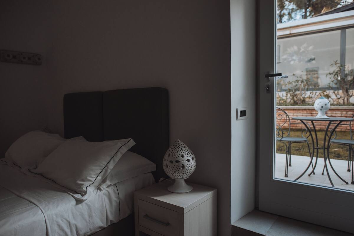 A bedroom with a bed, a bedside table with a lamp, and a view of an outdoor patio through a door.