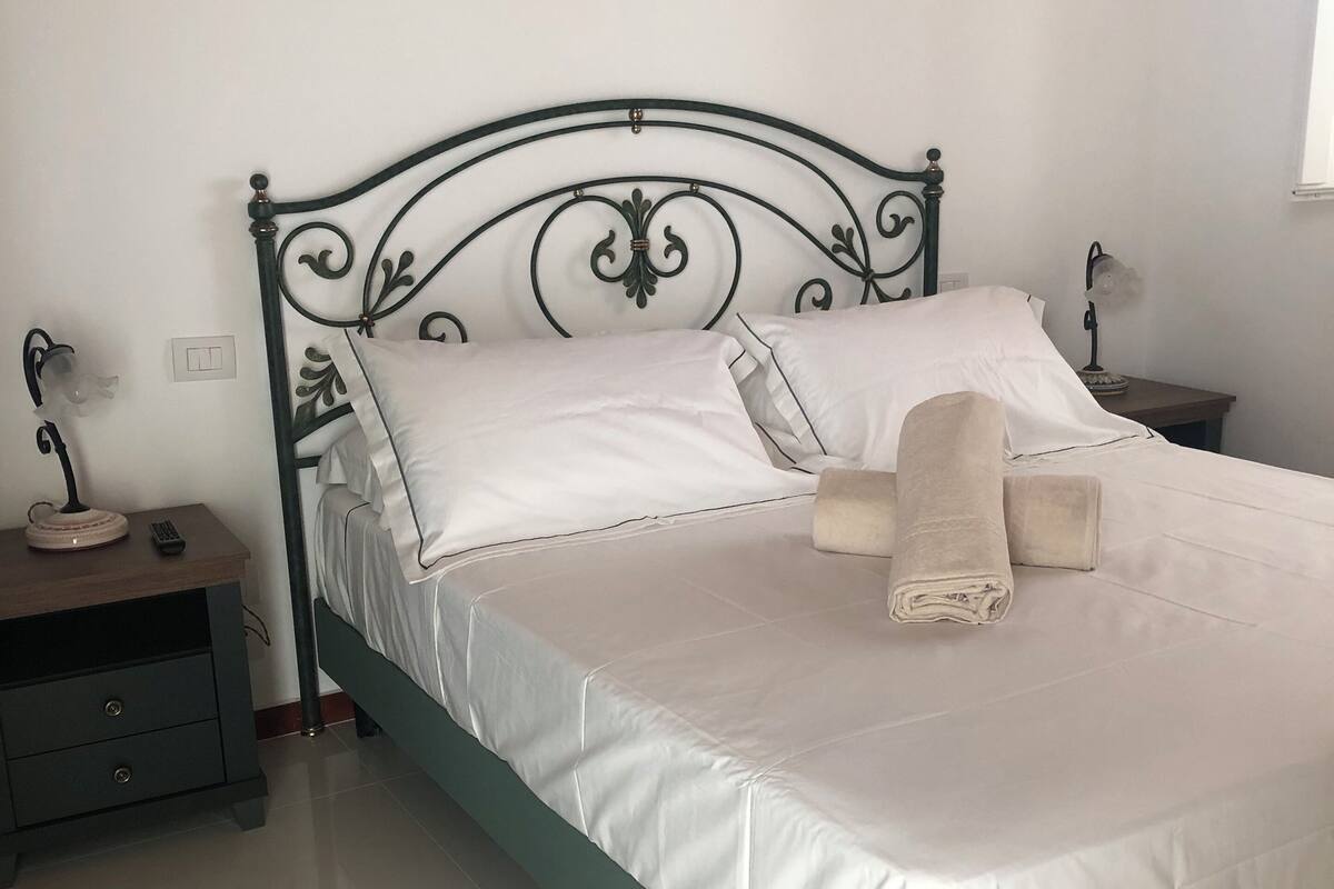 A neatly made bed with white linens and two folded towels, a bedside table with a lamp, and a nightstand with a remote control.