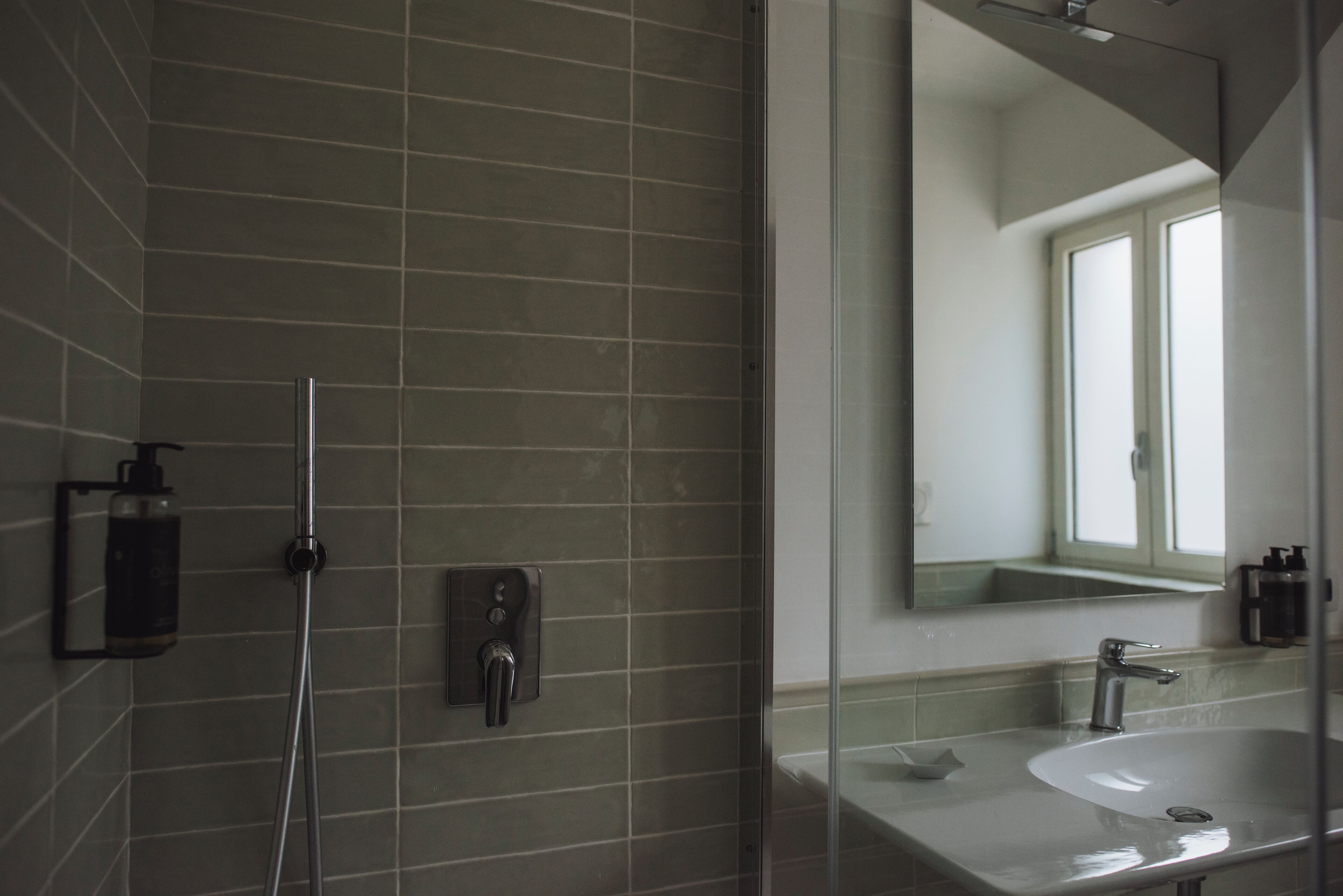 Un bagno moderno con cabina doccia in vetro, erogatore di sapone a parete e un ampio specchio che riflette una finestra.