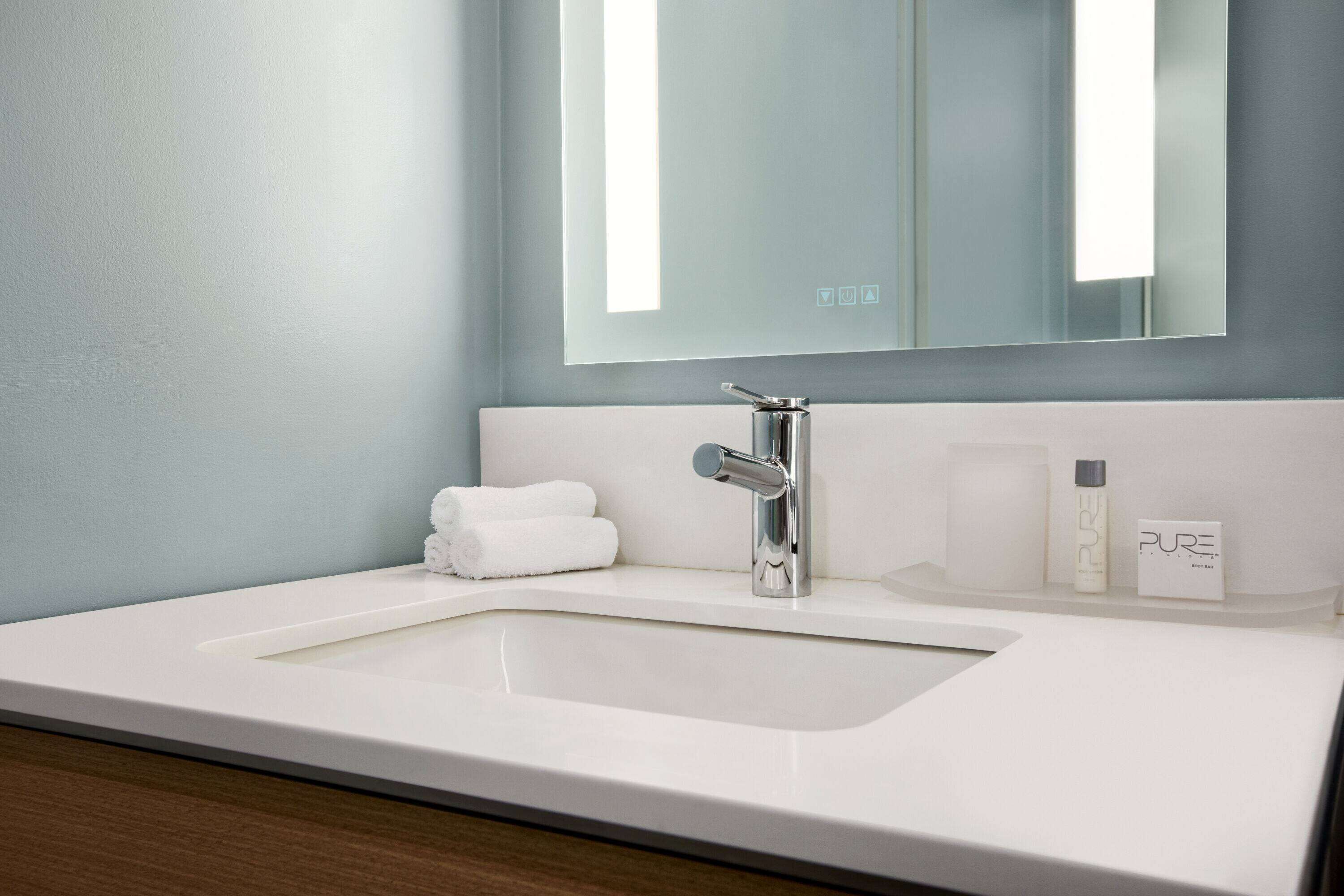 A modern bathroom with a white sink, a chrome faucet, and a mirror above it. There are two white towels and a bottle of Pure shampoo on the counter.