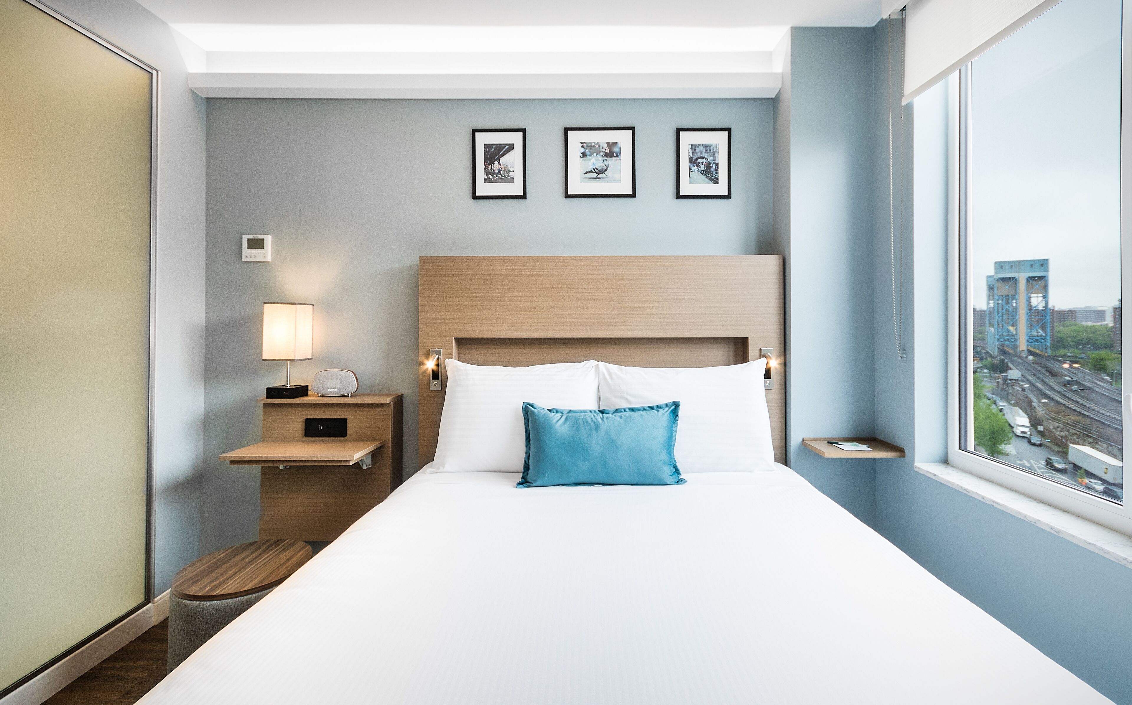 A hotel room with a bed, a nightstand with a lamp, a small table, and a window with a city view.