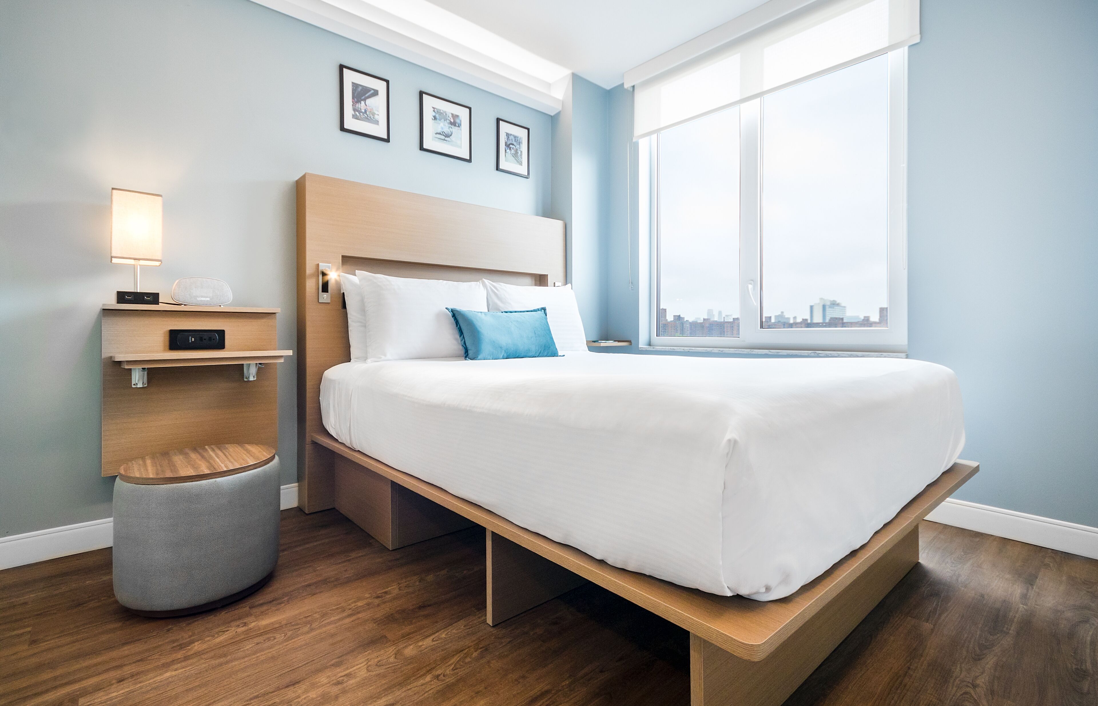 A modern hotel room with a wooden bed, a bedside table with a lamp, a small stool, and a large window offering a city view.