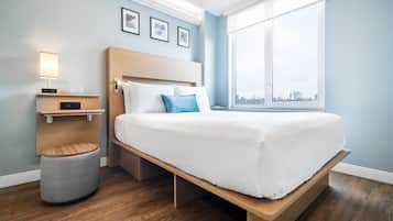 A modern hotel room with a wooden bed, a bedside table with a lamp, a small stool, and a large window offering a city view.
