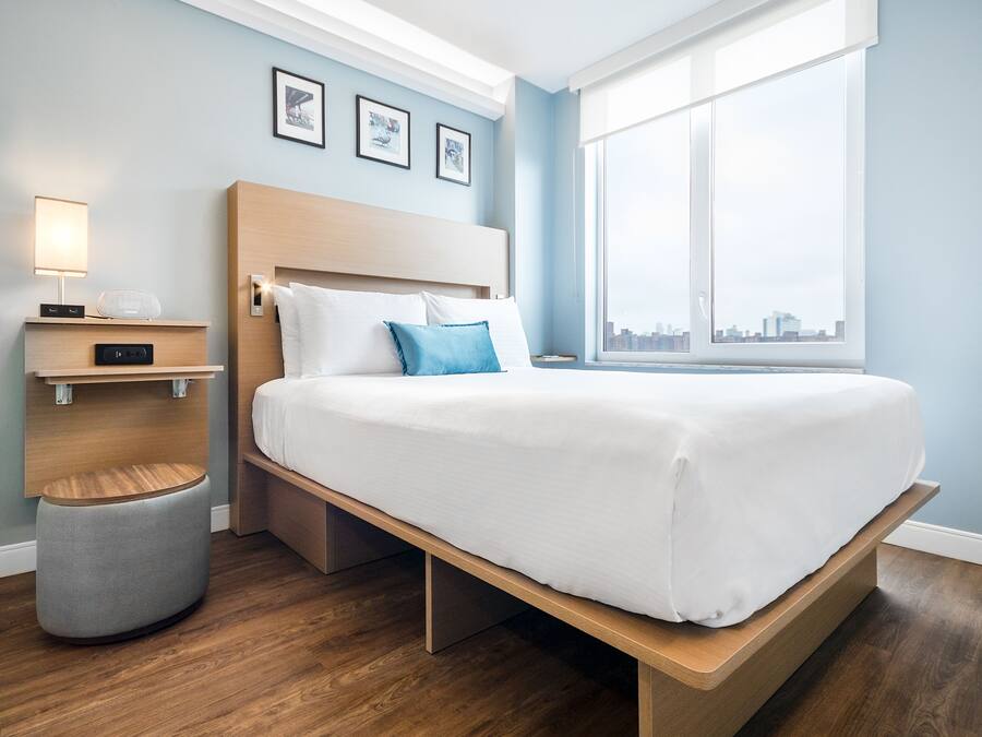 A modern hotel room with a wooden bed, a bedside table with a lamp, a small stool, and a large window offering a city view.
