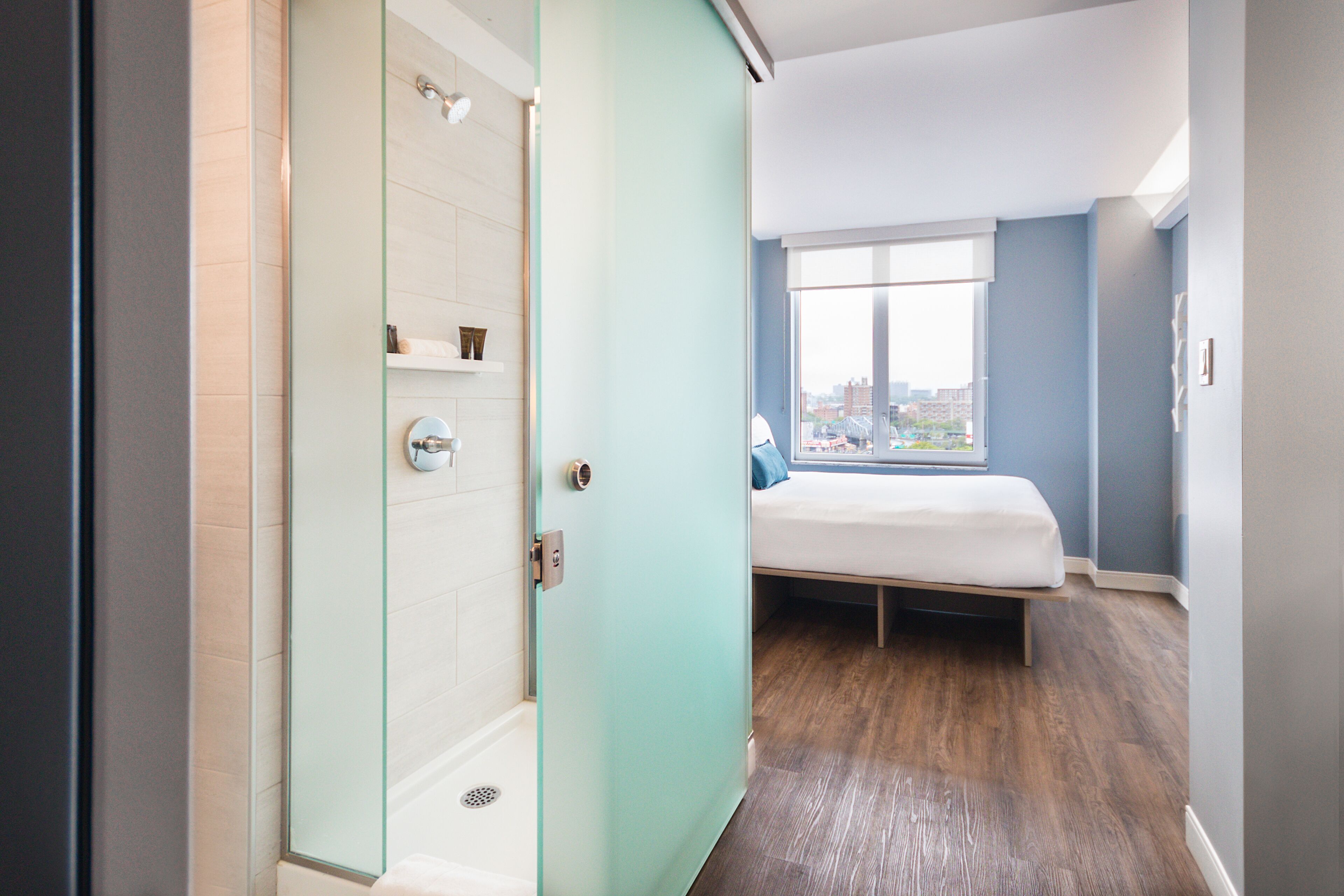 A modern hotel room with a glass shower enclosure, a bed with a city view, and a large window.