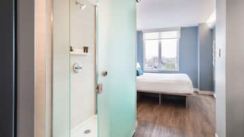 Ein modernes Hotelzimmer mit einer Duschkabine aus Glas, einem Bett mit Blick auf die Stadt und einem großen Fenster.