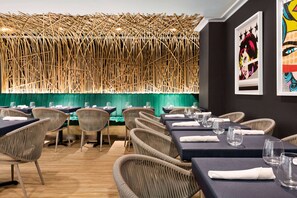 A restaurant with wicker chairs and tables, a green upholstered booth, and a bamboo wall.