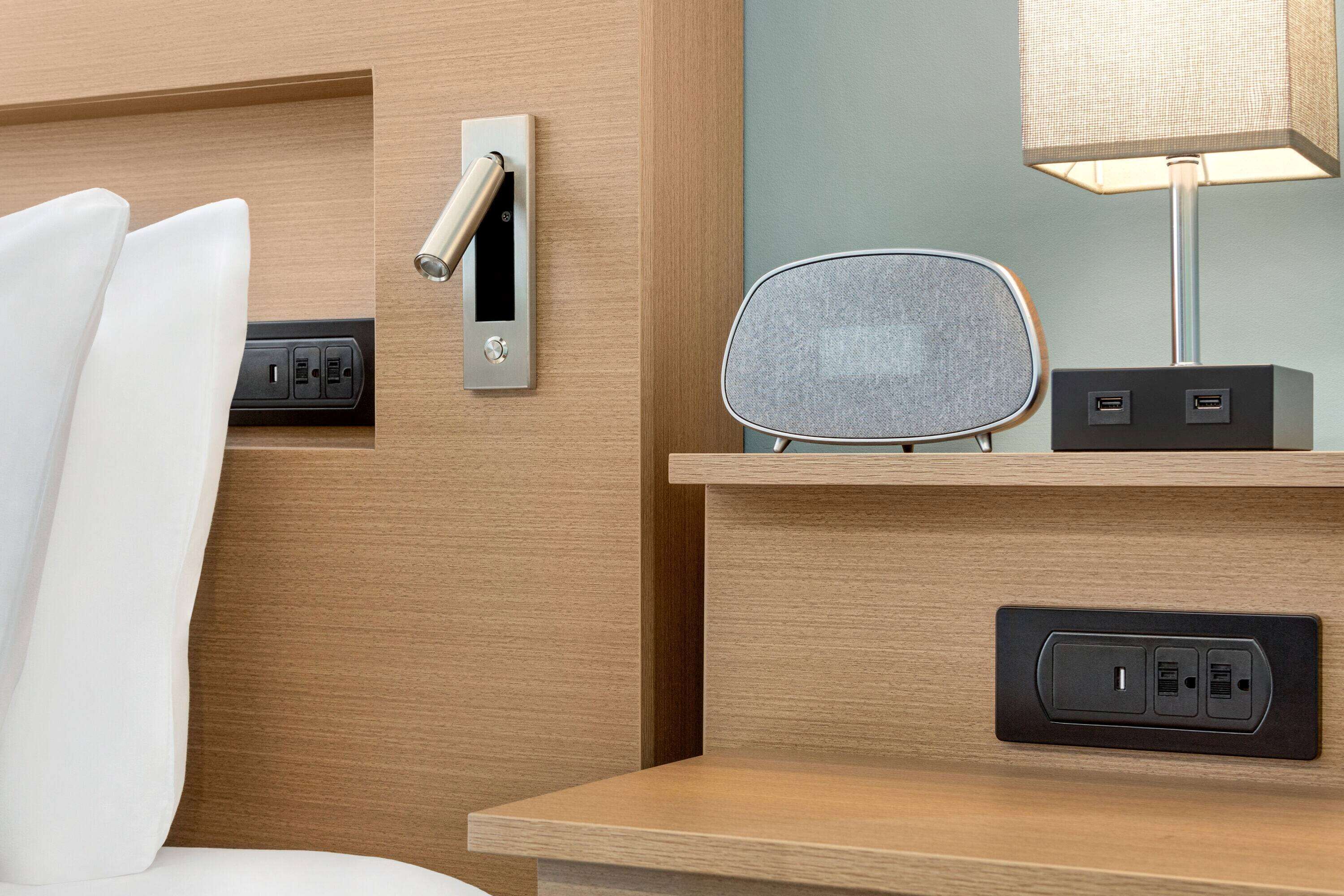 Habitación de hotel con cabecera de madera, mesita de noche con lámpara, toma de corriente y altavoz.