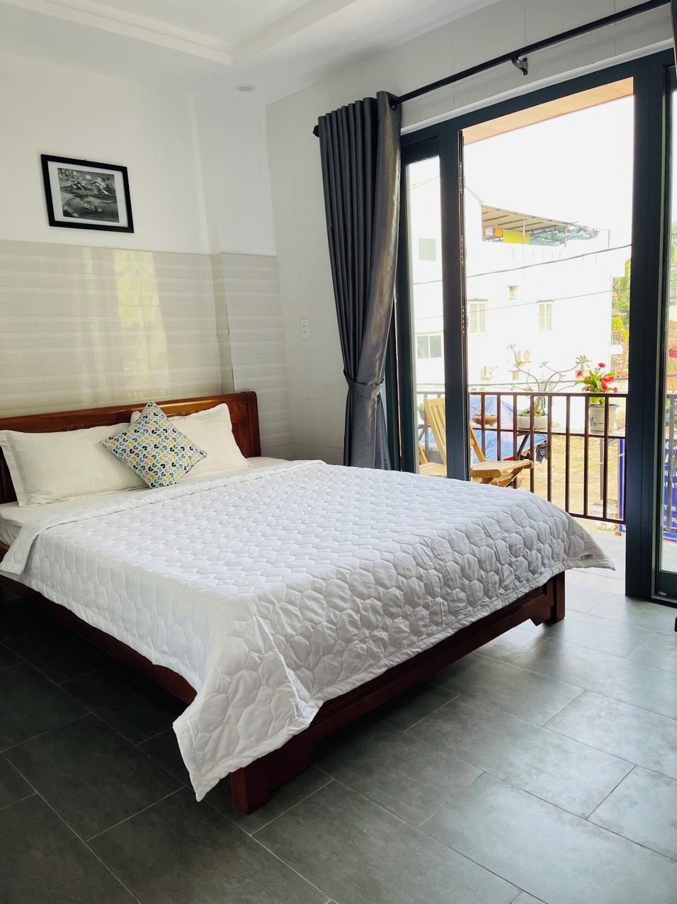 A bedroom with a bed, a window with curtains, a picture on the wall, and a view of the outside.