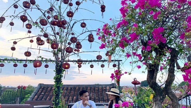 A dining area with tables and chairs, a person sitting at a table, and a tree with pink flowers. - Homestay Cuong Thinh (Da Nang)
