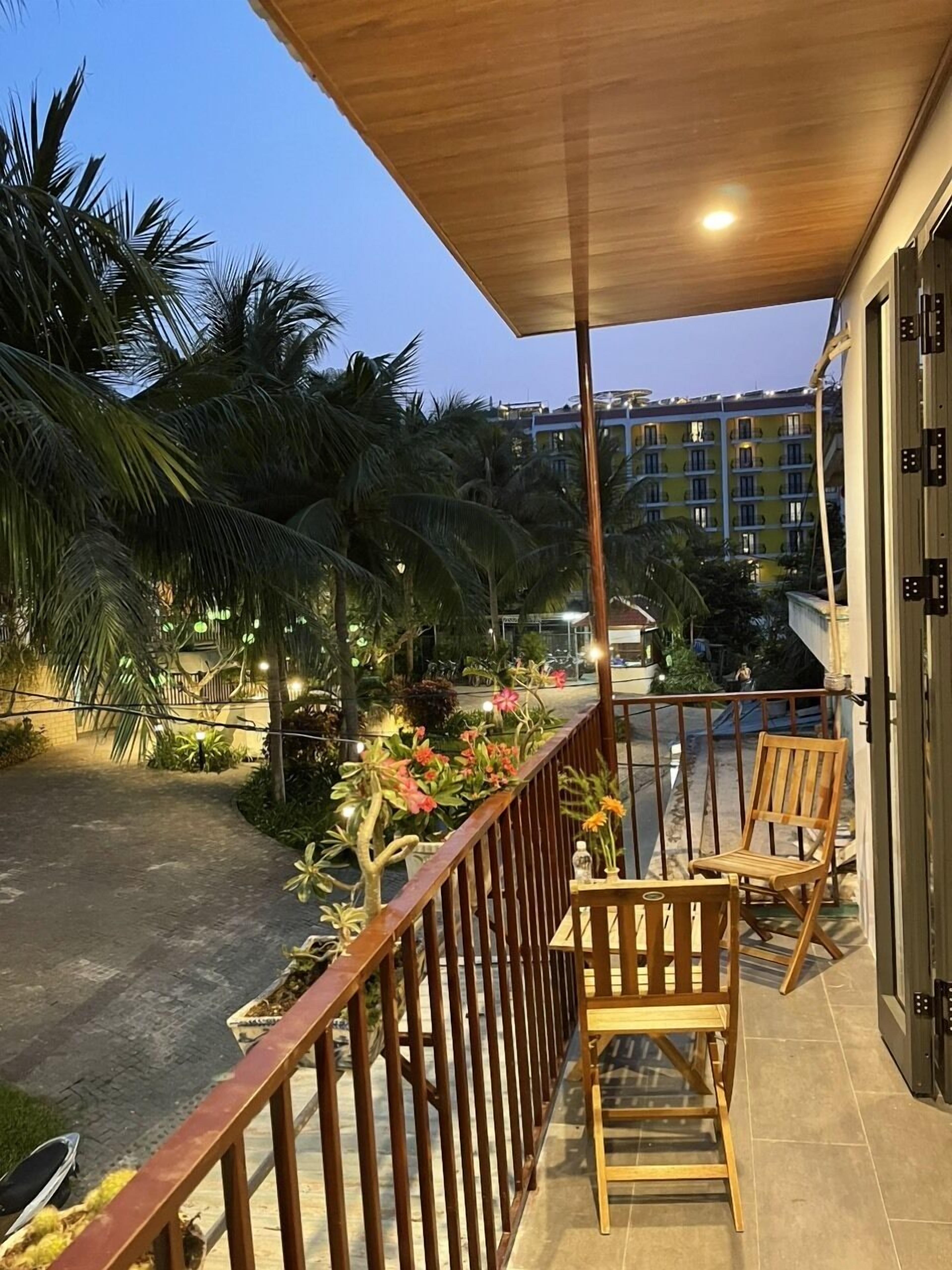 A balcony with wooden chairs, a railing, and a view of palm trees and a building in the background.