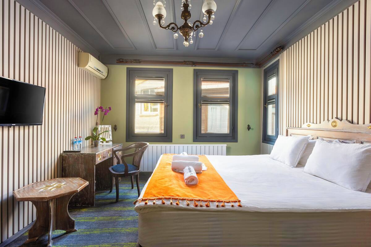 Habitación de hotel con una cama grande, un escritorio con silla, una televisión y dos ventanas con persianas.