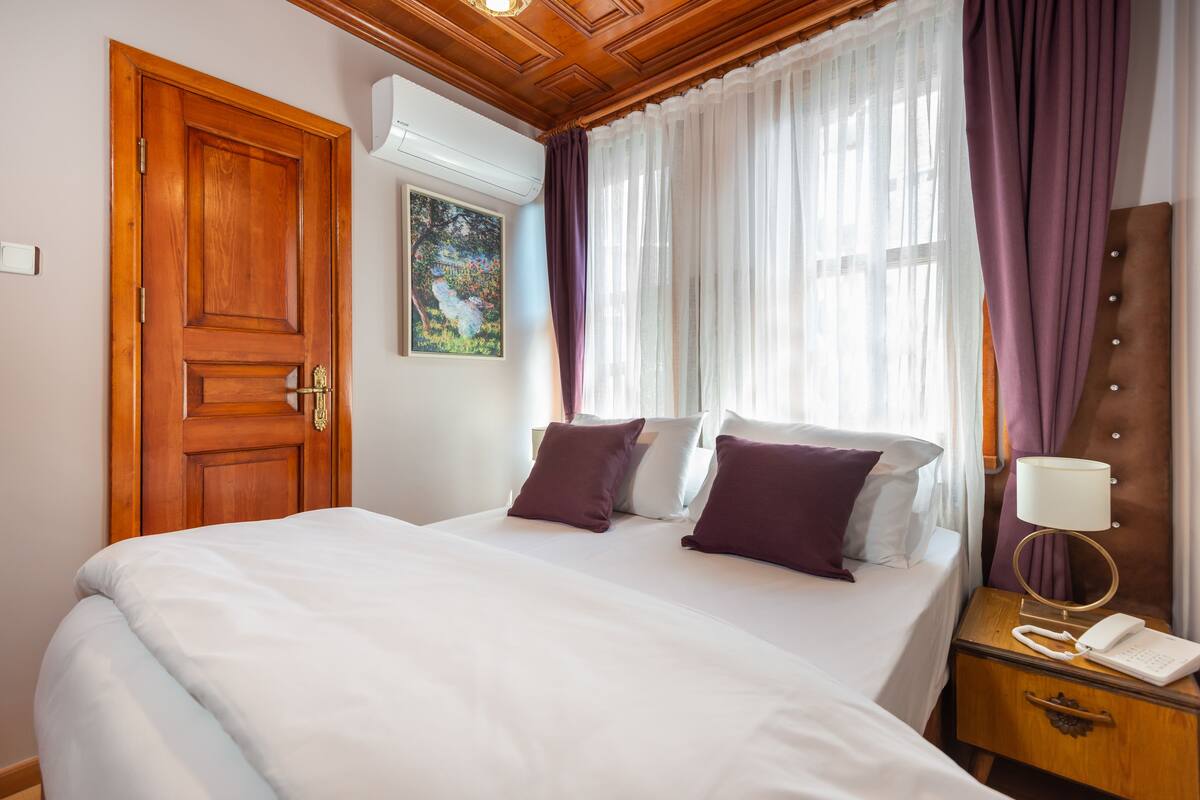 Habitación de hotel con puerta de madera, cama con ropa de cama blanca y morada, mesita de noche con lámpara y un cuadro en la pared.