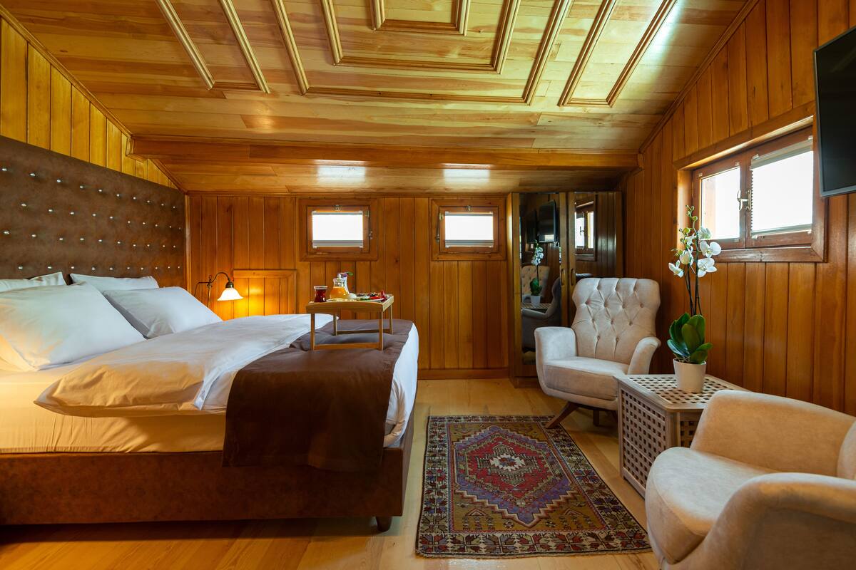 Una habitación con paneles de madera, una cama, una silla, una mesita y una planta en maceta.