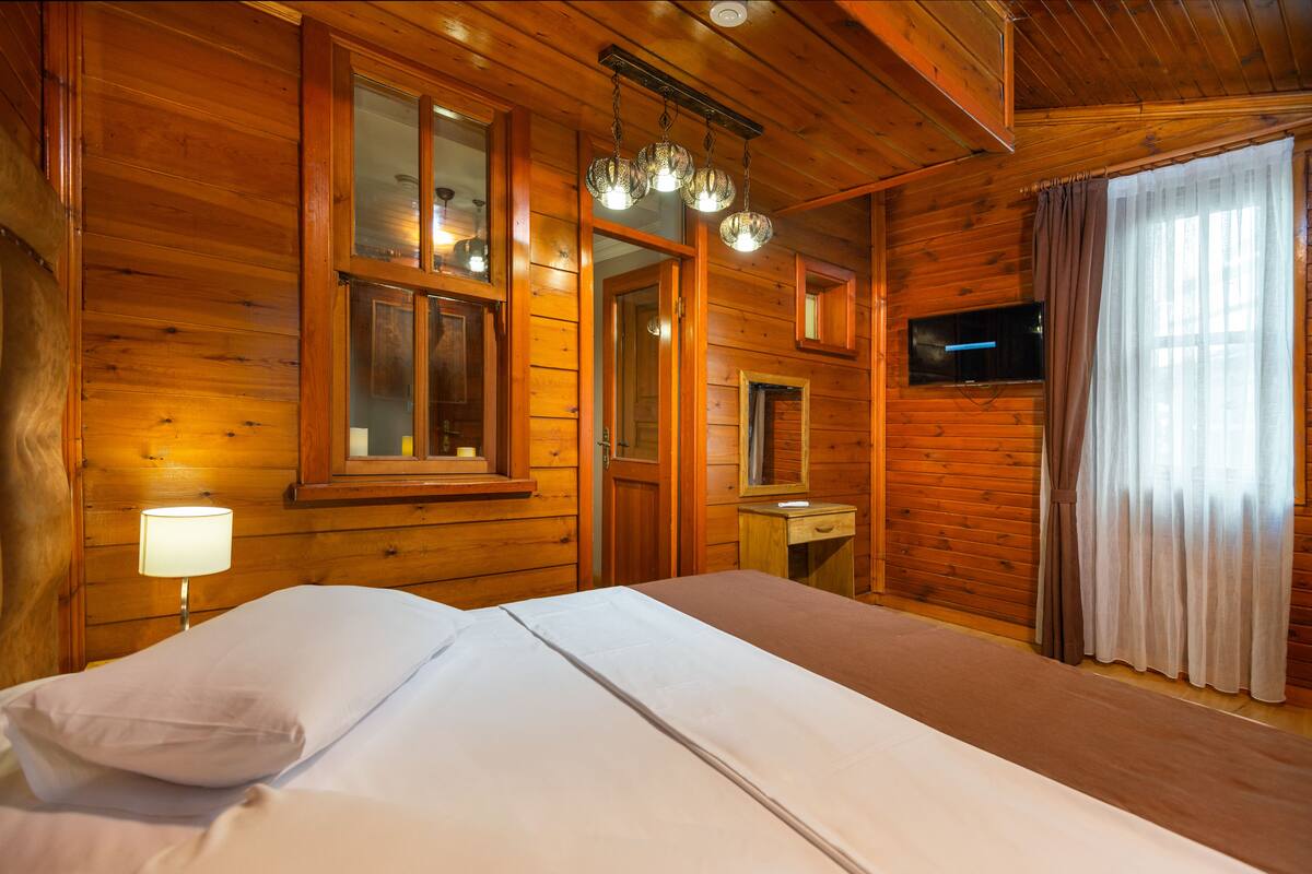 Habitación de cabaña de madera con cama, mesita de noche, lámpara, televisor y ventana con cortinas.