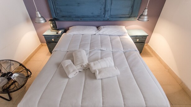 Un lit bien fait, avec du linge de lit blanc et des serviettes pliées, une table de chevet avec une lampe et un ventilateur posé sur le sol.